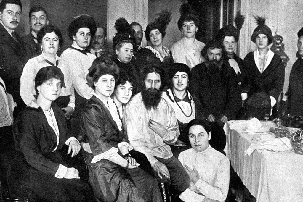Image of a black and white photo of Rasputin with his characteristic beard and intense gaze, sitting among well-dressed Russian aristocrats in an opulent setting.