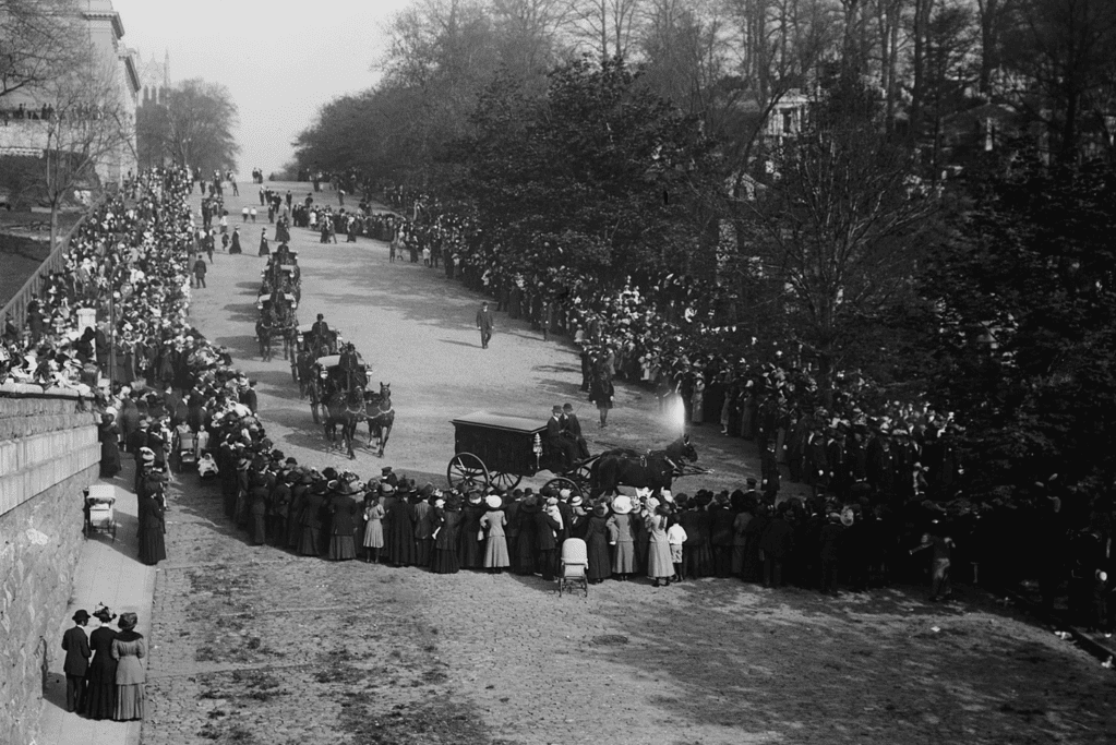Image of a photo of a funeral procession featuring horse-drawn carriages, including one bearing the casket of John Astor IV, in a moment of mourning and remembrance.