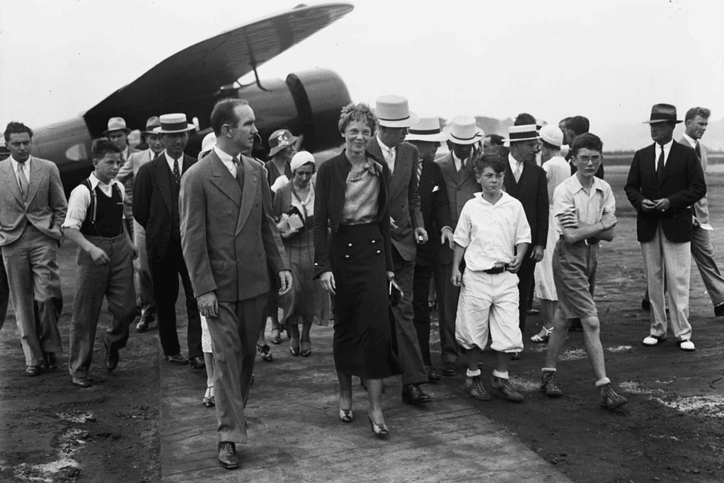 Image of Amelia Earhart, always the fasihon icon, walking with a crowd at an airfield.